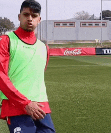 a man in a green vest is standing on a soccer field with a coca cola banner in the background .