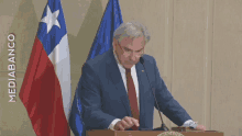 a man in a suit and tie stands at a podium in front of two flags and the words mediabanco on the wall behind him