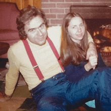 a man wearing suspenders sits next to a woman in blue pants