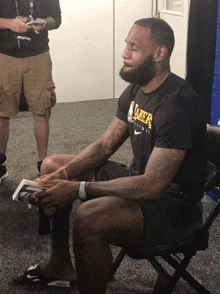 a man wearing a black shirt that says lakers sits in a chair