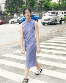 a woman wearing a purple dress and black slides with slim on them crosses a crosswalk