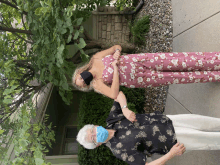 a woman in a pink floral jumpsuit holds the hand of an older woman wearing a blue mask