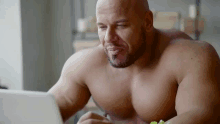 a shirtless man is sitting at a table with a laptop and eating a salad .