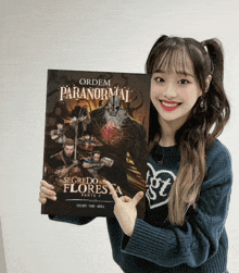 a girl in a blue sweater is holding a book called ordem paranormal