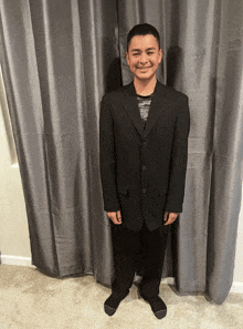 a young man wearing a black suit and black socks smiles in front of a grey curtain