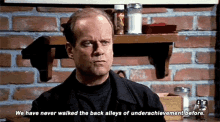 a bald man is standing in front of a brick wall and a shelf with salt and pepper shakers .