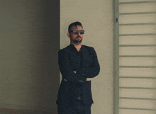 a man in a suit and sunglasses stands with his arms crossed in front of a wall