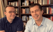 two men are sitting in front of a bookshelf and one of them is wearing glasses and a blue shirt