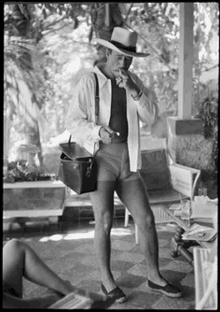 a man wearing a hat and shorts is standing on a porch smoking a cigarette