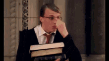 a man wearing glasses and a tie is holding a stack of books and rubbing his nose .