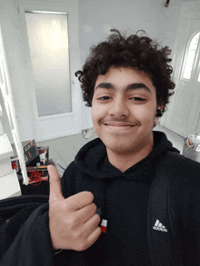 a young man with curly hair wearing an adidas hoodie gives a thumbs up