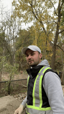 a man wearing a safety vest and a hat stands in front of a forest