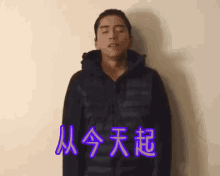 a man in a black jacket is standing with his eyes closed in front of a wall with chinese writing on it .