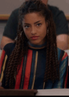 a woman with dreadlocks is sitting at a table in a classroom wearing a striped shirt and choker .