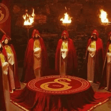 a group of people in red cloaks are standing around a round table with candles .