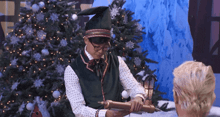 a man dressed as a christmas elf holds a scroll in front of a christmas tree