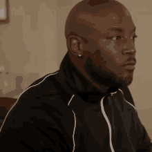 a bald man with a beard and earrings is wearing a black jacket and sitting at a table .