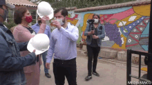 a man wearing a mask holds a white hard hat in front of a mural that says mediabanco on the bottom