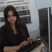 a woman is typing on a keyboard in front of a monitor with a newspaper on the wall behind her