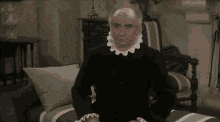 an elderly man is sitting on a couch in a living room wearing a black dress and a white collar .