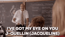 a man in a suit and tie is standing in front of a blackboard in a classroom talking to a group of students .