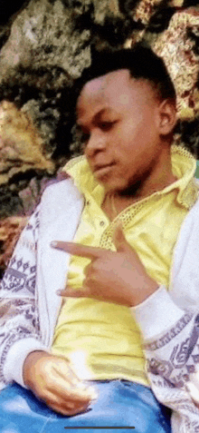 a young man wearing a yellow shirt and a white jacket is sitting on a rock and making a funny face .