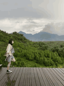 a woman wearing a mask is standing on a wooden deck overlooking a lush green forest