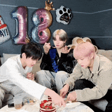 three young men are sitting on a couch eating cake with balloons in the background that say happy 18th birthday