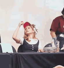 a woman wearing a baseball cap is sitting at a table with a stack of plates and a bottle of water .