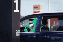 a green cartoon character is sitting in a car next to a place order here sign