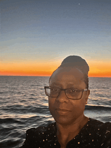 a woman with glasses stands in front of the ocean at sunset