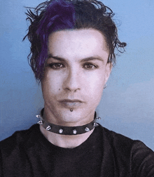 a man with purple hair and a black choker looks at the camera