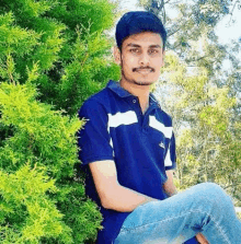 a young man is sitting in front of a tree wearing a blue polo shirt and jeans .