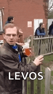 a man is holding a glass of beer and smoking a cigarette while standing next to a wooden fence .