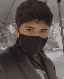 a young man wearing a black mask and a black jacket stands under an umbrella