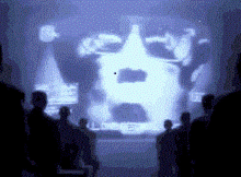 a group of people are standing in front of a large screen with a face projected on it