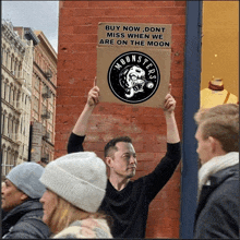 a man holds up a sign that says " buy now don t miss when we are on the moon "