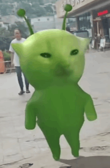 a green alien with a cat 's head and ears is walking down a sidewalk .