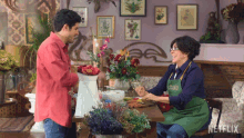 a man in a red shirt talks to a woman in a green apron who is sitting at a table