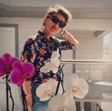 a woman wearing sunglasses and a floral shirt stands in front of flowers