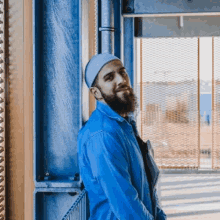 a man with a beard wearing a hat and a blue jacket is leaning against a blue wall .