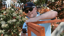 a man wearing sunglasses and a baseball cap is smoking a cigarette while holding a drill .