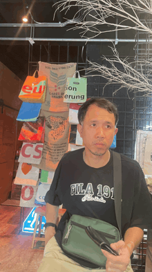 a man wearing a fila 191 t-shirt stands in front of a wall of bags
