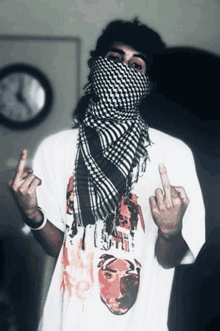 a man wearing a scarf around his face and a t-shirt with tupac shakur on it is giving the middle finger .