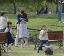 a man is hugging a woman in a park while a group of people are standing around a bench .