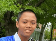 a young boy with a shaved head is smiling for the camera in front of a tree .