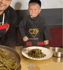 a little boy is sitting at a table with a plate of food and a spoon in his hand .