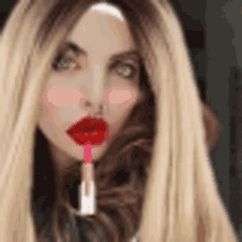 a woman with long blonde hair and red lipstick on her lips is holding a lipstick in her mouth .