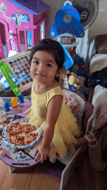 a little girl in a yellow dress stands in front of a frozen table with a plate of pizza on it