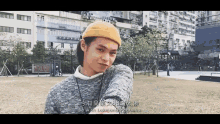 a young man wearing a yellow hat and a grey sweater is standing in a park with buildings in the background